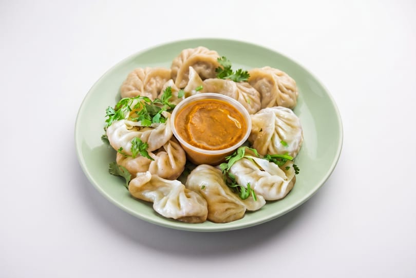 A light green plate filled with steamed momos, garnished with cilantro, and a cup of dipping sauce.