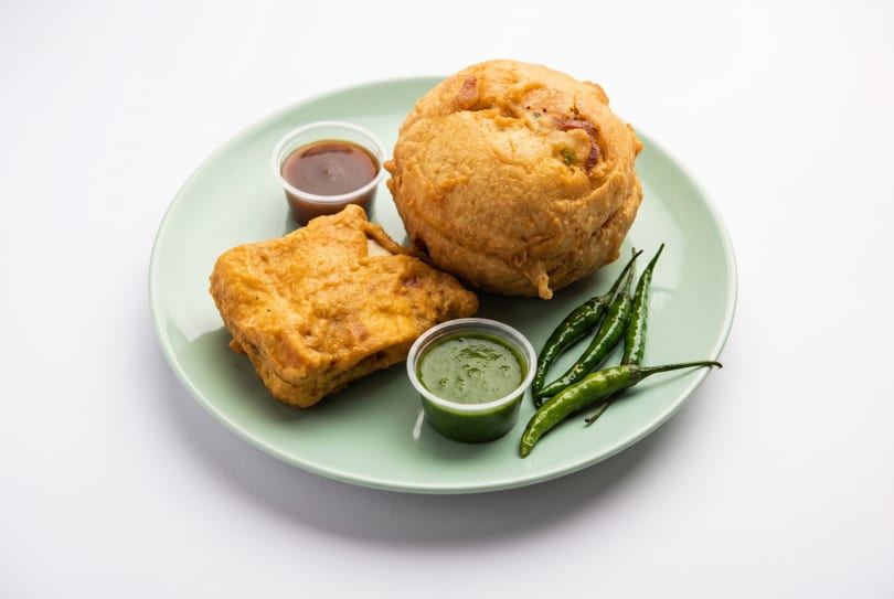 Fried Indian snacks, including a vada pav and a batata vada, with chutneys and green chilies.