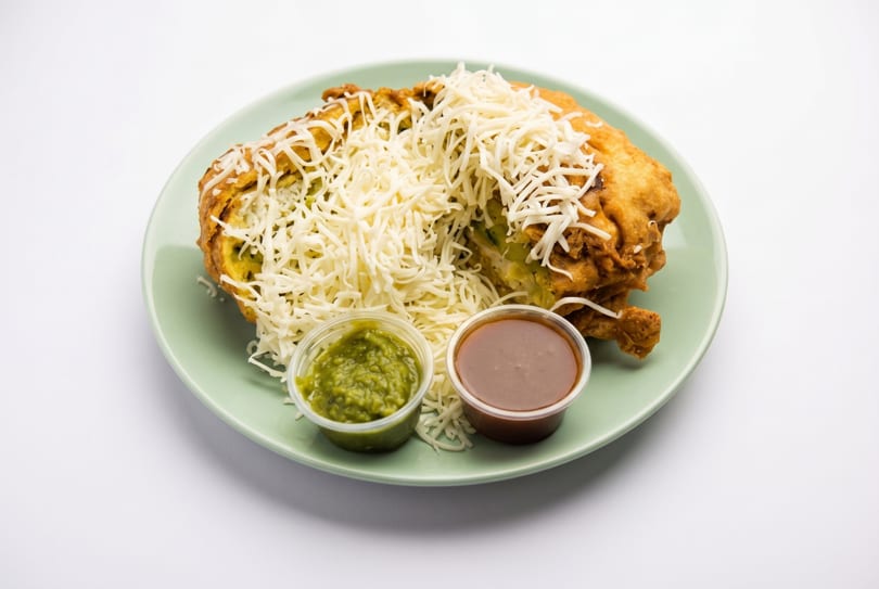 Deep-fried Indian snack, Vada Pav, topped with shredded cheese and served with green and brown chutneys.