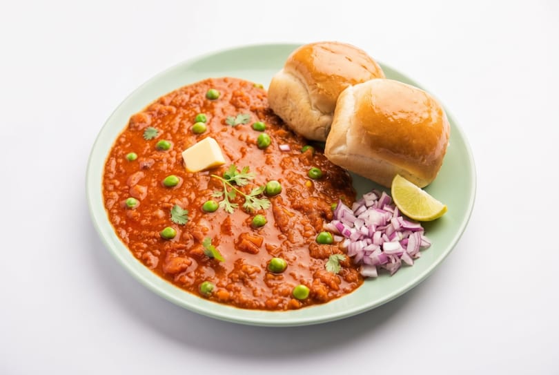 Pav Bhaji, a vibrant red vegetable curry with green peas, butter, buns, chopped onions, and lime.
