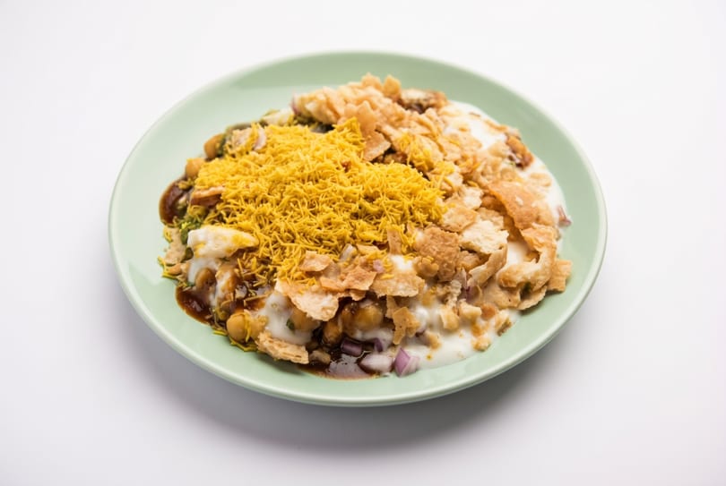 A vibrant Indian chaat dish with yogurt, chutneys, chickpeas, sev, and crispy flatbread on a green plate.